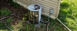Dirty outdoor AC condenser unit clogged with branches and overgrowth at the end of summer in Malvern, PA, needing repair and cleaning.