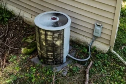 Dirty outdoor AC condenser unit clogged with branches and overgrowth at the end of summer in Malvern, PA, needing repair and cleaning.