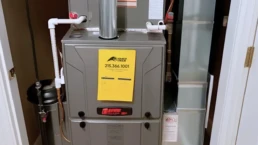 Gas furnace in a utility closet with a yellow maintenance tag on the front panel and PVC exhaust pipes above it.