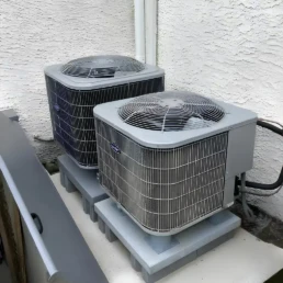 Two outdoor air conditioning condenser units on a concrete pad beside a textured stucco wall.